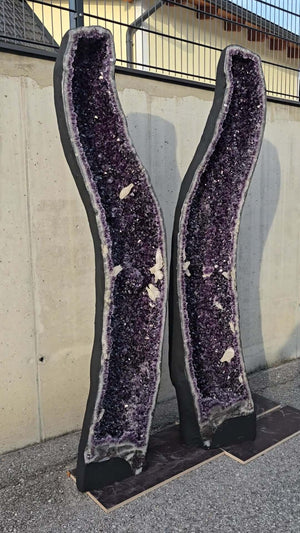 Pair of tall, dark purple amethyst geode halves displayed against a concrete wall, showcasing intense color and crystal quality.
