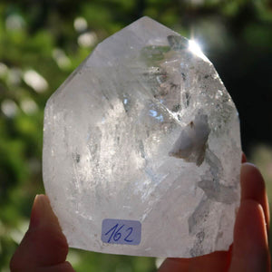 Hand holding polished Bergkristall quartz crystal point, Unikat Nr. 162, shining with faceted surfaces in natural sunlight, Brazil.