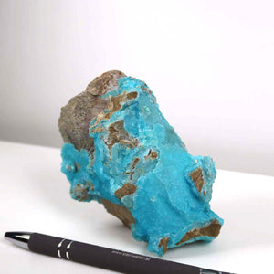 Turquoise-blue Hemimorphite rough stone with pen for scale, showcasing natural mineral color and texture.