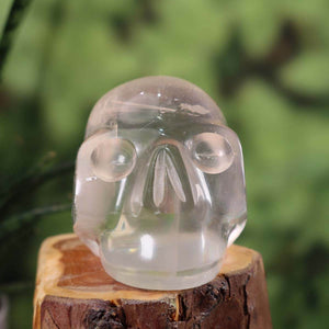 Handcrafted white calcite crystal skull with unique shape, 7x7 cm, displayed on wooden base against green background.