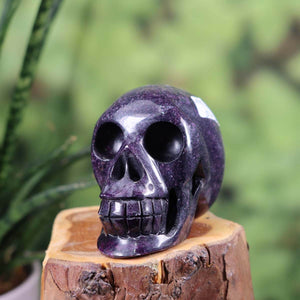 Handcrafted crystal skull made from natural stone, 7x6 cm, displayed on a wooden stand against a blurred green background.
