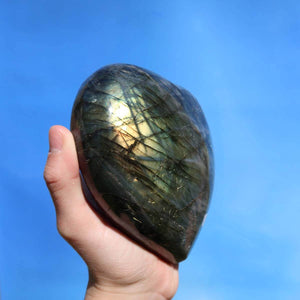 Hand holding a large Labradorite heart stone reflecting colors in sunlight, showcasing unique patterns, used for relaxation and creativity.
