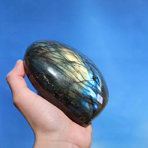 Hand holding large labradorite heart gemstone with blue and gold hues against a blue sky