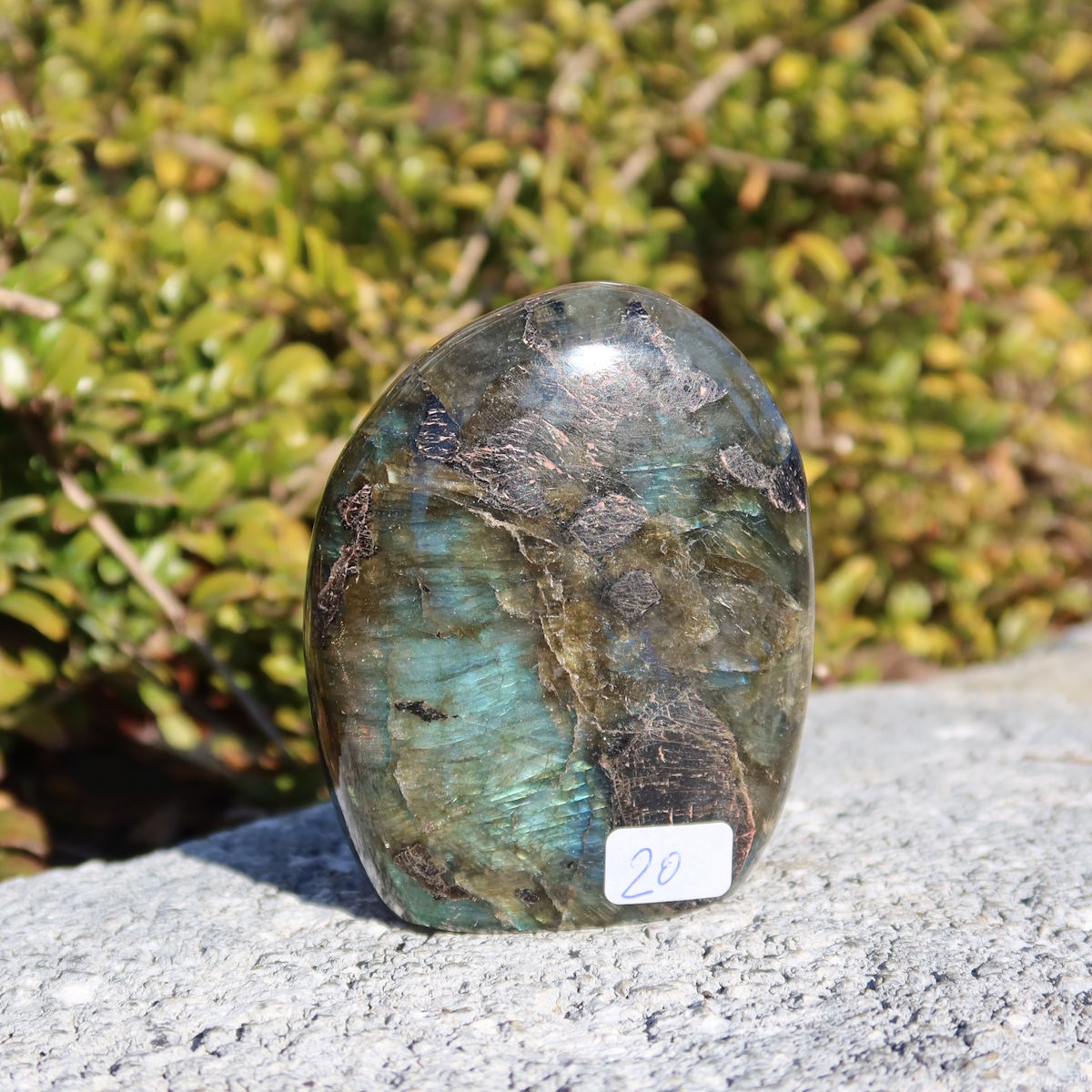 Labradorit Freiform poliert - 7x5cm - Intuition und Kreativität Nr.20 auf grauem Stein hinter grünem Hintergrund.
