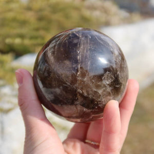 Hand holding polished smoky quartz sphere with mystic brown-gray hues; buy at Steinwelten Graz and Wiener Neustadt Kristallschädel shop.