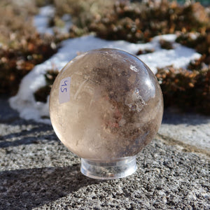 Rauchquarz Kugel – Natürliche Kristallkugel 60mm Durchmesser Nr. 45 auf einem Stein im Freien bei Sonnenlicht