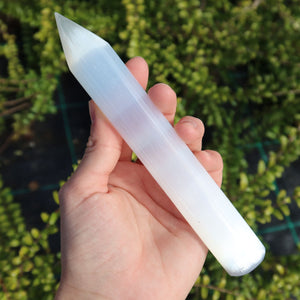 Selenit Massage Griffel mit Spitze- Akupunktur-Massagestab - 15cm in der Hand gehalten, vor Grünpflanzen.