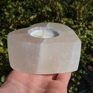 Hand holding a heart-shaped selenite tealight holder, polished and smooth, with natural mineral texture, against a green background.