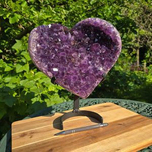 Amethyst Druse Amethyst Geode in Herzform mit Metallsockel 32x28x14cm - NR-23 auf einem Holztisch im Freien.