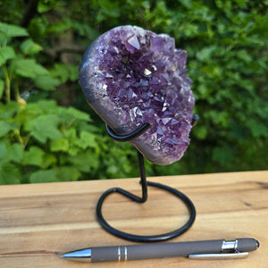 Amethyst Druse Amethyst Geode in Herzform mit Metallsockel 16x11x10cm - NR-61 auf einem Ständer, violette Kristalle vor grünem Hintergrund.