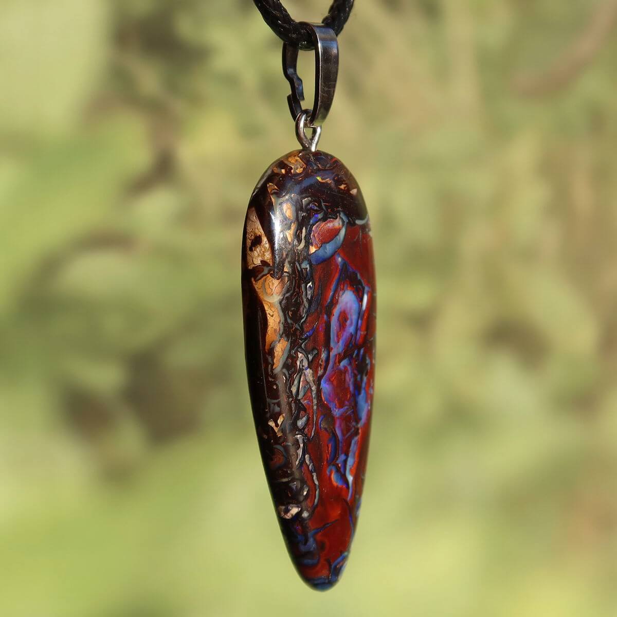 Boulder Opal Anhänger - Positivität und Freude - Nr. 35 mit einzigartigen Mustern auf silberner Öse vor grünem Hintergrund.