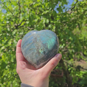 Hand holding a large Labradorite heart stone with blue and green hues, 9x9x4cm, against a lush green background, Unikat Nr. 9.