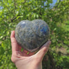 Hand holding a large labradorite heart with vibrant colors and unique patterns, set against a green natural background.