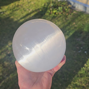 Hand holding a 14cm Selenite crystal sphere in sunlight, promoting light and protection, available for online purchase in Graz and Wiener Neustadt.