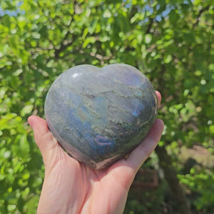 Hand holding large Labradorite heart gemstone with blue, green, and gold hues, unique piece for relaxation and creativity enhancement.