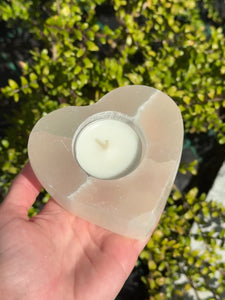 Heart-shaped selenite tealight holder with candle, polished natural mineral from Morocco, held against a leafy background.