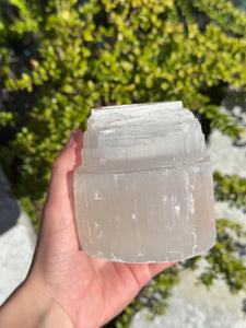 Hand holding a natural selenite crystal tealight, known as a light bringer and protective stone, against a green garden backdrop.