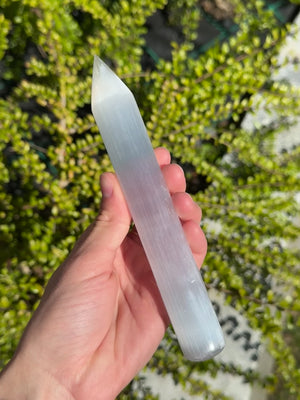 Selenit Massage Griffel mit Spitze- Akupunktur-Massagestab - 15cm in der Hand mit grünen Pflanzen im Hintergrund.